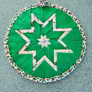 May include: A green and white fabric pot holder with a holly border. The pot holder is shaped like a star and has a loop for hanging.