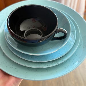 May include: A black teacup with a handle sits atop a stack of four light blue plates. The plates have a speckled pattern. The teacup and plates are held in a person's hand.