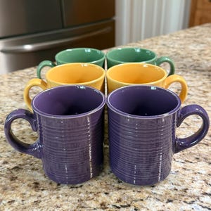 May include: A set of six ceramic mugs in various colors: purple, yellow, green, and orange. Each mug has a ribbed texture and a curved handle. The mugs are arranged on a speckled countertop.