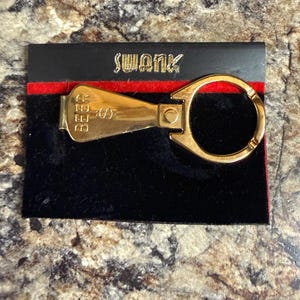 Op de afbeelding: Een goudkleurige sleutelhanger met een scharnierend ontwerp, met het woord "SWANK" in gouden letters op een zwarte achtergrond met een rode streep. De sleutelhanger ligt op een zwart fluwelen oppervlak.