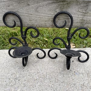 May include: A pair of black metal candle holders featuring a scrollwork design with multiple curved accents and a circular base for holding a candle. The structure is composed of dark, matte-finished metal wire shaped into decorative, swirling patterns.