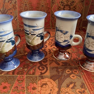 May include: Four ceramic mugs with blue and white seaside designs. Each mug has a different design featuring a lighthouse, seagulls, and seashells. The mugs have a brown base and a blue and white design.