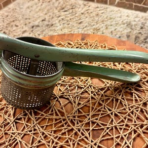 Puede incluir: Un pasapurés antiguo con acabado verde claro. El utensilio de cocina tiene un cuenco de metal perforado y mangos largos para prensar patatas. El pasapurés está sobre un salvamanteles tejido y una tabla de cortar de madera.