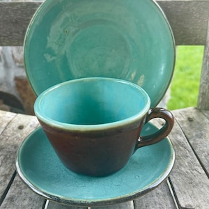 May include: A teal ceramic teacup and saucer set with a brown exterior and a teal interior. A matching teal plate is behind the teacup. The set is on a weathered wooden surface.