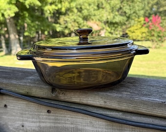 May include: A vintage, amber-colored glass casserole dish with a matching lid. The dish has a round shape with two handles and a knob on the lid. The dish is sitting on a wooden surface, with a blurred green background.