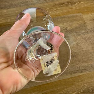 May include: A clear, stemmed glass with a rounded bowl and a wide base. The glass has a subtle iridescent sheen, reflecting colors. A partially visible sticker is affixed to the base. The glass is held in a person's hand.