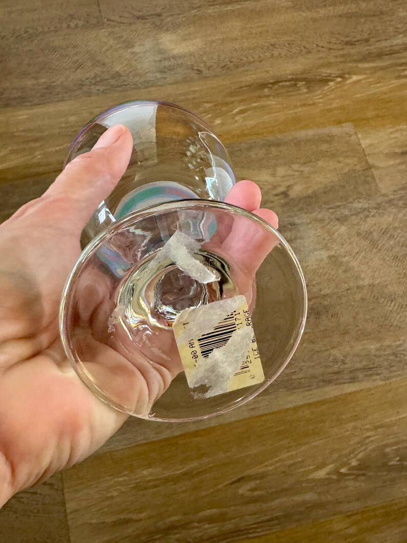 May include: A clear, stemmed glass with a rounded bowl and a wide base. The glass has a subtle iridescent sheen, reflecting colors. A partially visible sticker is affixed to the base. The glass is held in a person's hand.