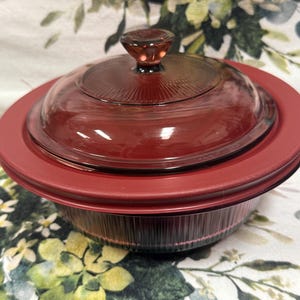 Può includere: Una pirofila in vetro rosso con coperchio abbinato e bordo rosso. Il coperchio ha una piccola maniglia arrotondata. Il piatto è appoggiato su una superficie a motivi floreali.