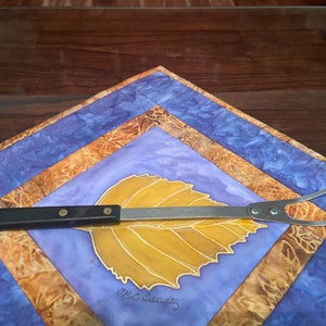 May include: A stainless steel grilling fork with a black handle, featuring a leaf design on a blue and gold fabric background.