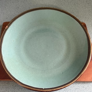 Può includere: Un piatto rotondo in ceramica verde chiaro con un bordo marrone scuro. Il piatto è appoggiato su una piccola superficie di legno rettangolare. L'interno del piatto presenta un sottile disegno a cerchi concentrici.
