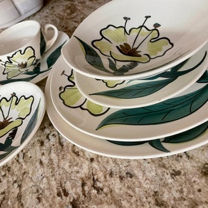May include: A set of four white ceramic plates with a green and yellow floral design. The plates are stacked on top of each other, with the largest plate on the bottom and the smallest plate on top. The plates feature a stylized floral design with green leaves and yellow flowers.