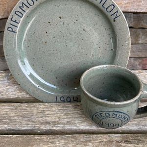 May include: A speckled sage green ceramic plate and matching mug set. The plate features the text PIEDMONT TRIATHLON along the rim and 1994 at the bottom. The mug displays a circular stamp with the text PIEDMONT 1994 on the front surface.