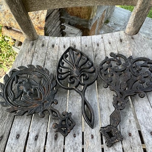 May include: Three black cast iron trivets with intricate designs. The trivets are arranged on a wooden surface.