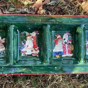 Puede incluir: Una bandeja de servir de cerámica verde con cuatro secciones divididas. Cada sección presenta una escena navideña festiva con Santa Claus y otros personajes. La bandeja tiene un borde rojo y un glaseado blanco moteado.