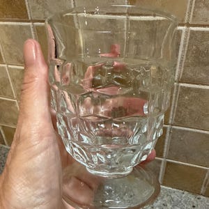 May include: A clear, vintage-style glass with a textured, geometric pattern around the bowl and a rounded base. The glass is held in a hand, showcasing its transparency and design. Ideal for serving beverages.