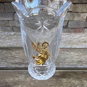 Op de afbeelding: Een heldere glazen vaas met een gegolfde rand en een diamantpatroon. Een gouden engelbeeld is aan de voorkant van de vaas bevestigd.