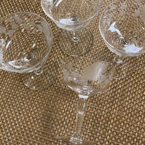 May include: A set of four clear glass coupes featuring etched floral and leaf patterns around the rim. Each glass has a wide, shallow bowl, a slender stem, and a circular base. The etched designs include detailed flower petals and small leaves.