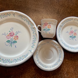 May include: A set of white ceramic dishes with a floral pattern in shades of pink, blue, and green. The set includes a large dinner plate, a smaller salad plate, a saucer, and a teacup.