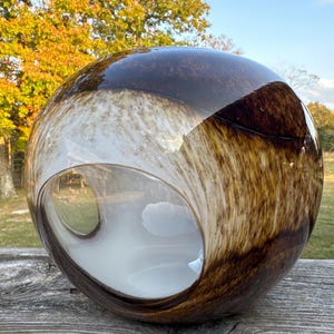 Puede incluir: Una gran esfera de vidrio marrón y blanco con una abertura circular. La esfera tiene una superficie moteada y texturizada.