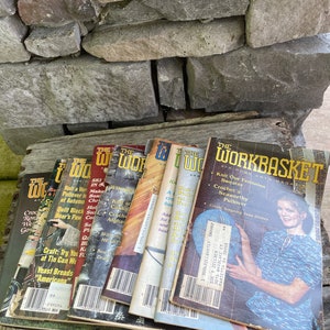 Puede incluir: Una pila de ocho revistas vintage de The Workbasket que presentan varios proyectos de artesanía, incluyendo tejido, ganchillo y acolchado. Las revistas tienen portadas coloridas con imágenes de proyectos terminados y texto que dice "The Workbasket" y "Knit Our Feminine Sweater".