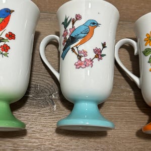 May include: Set of three white ceramic mugs with colorful bird and floral designs. Each mug has a different colored bird and a green, blue, or orange base.
