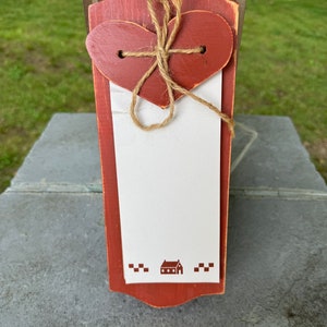 May include: A red wooden memo board with a heart-shaped cutout and a twine bow. The board has a white space for writing notes and a small house design at the bottom.