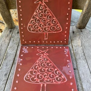 以下が含まれることがあります： クリスマスツリーの白い浮き彫りのデザインが施された赤いセラミックタイル2枚。それぞれのツリーは頂上に星があり、渦巻き模様で飾られています。