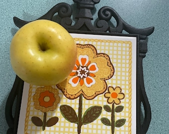 May include: A black cast iron trivet with a floral design. The trivet has a yellow, orange, and brown floral pattern on a white background. The trivet has a decorative handle on top.