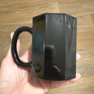 May include: A black ceramic mug with a unique octagonal shape and a handle. The mug is smooth and glossy.
