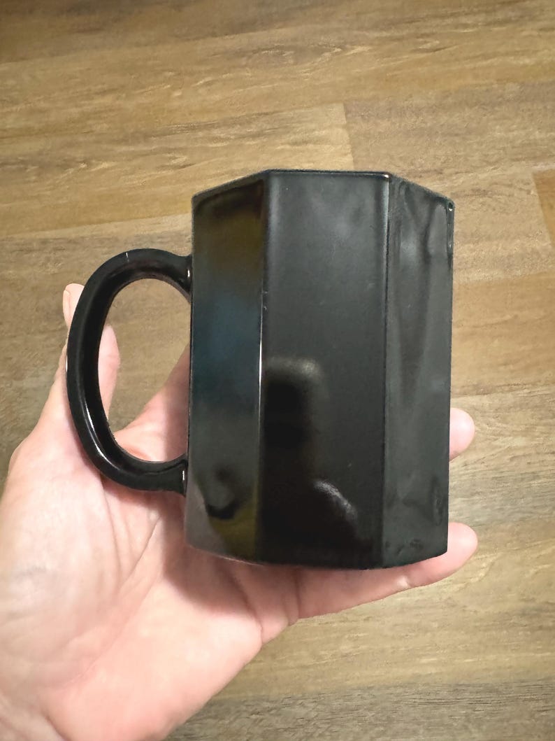 May include: A black ceramic mug with a unique octagonal shape and a handle. The mug is smooth and glossy.