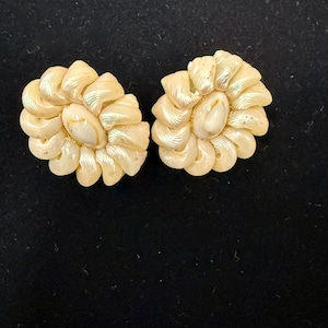 Op de afbeelding: Een paar crèmekleurige, bloemvormige oorbellen. Elke oorbel heeft een centraal ovaal element omgeven door gelaagde, bloembladachtige structuren. De oorbellen hebben een parelmoerglans.