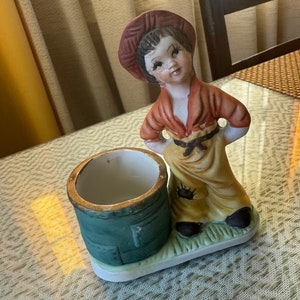May include: A porcelain figurine of a boy wearing a red hat and brown shirt, holding a green and white ceramic cup. The figurine is standing on a white base with green grass.