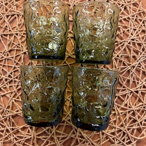 May include: Four olive green textured drinking glasses. The glasses have a unique, organic pattern and are arranged on a woven placemat. The glasses are cylindrical and appear to be made of glass.