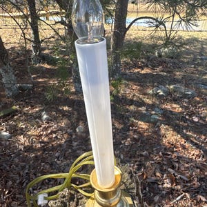 May include: A white electric candle with a clear glass bulb on top, sitting on a gold metal candlestick base. The candle is plugged into a cord.
