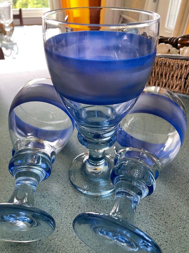 K&ouml;nnte beinhalten: Drei klare Glaskelche mit blauen, bemalten B&auml;ndern um den oberen und unteren Rand der Schale. Die Kelche haben einen dicken, klaren Glasstiel und einen breiten, klaren Glasfu&szlig;.