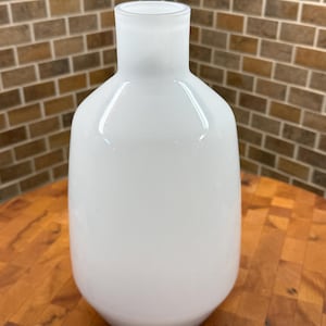 May include: A white glass vase with a rounded body and a narrow neck. The vase has a smooth, glossy finish and a frosted rim. The background features a brick wall and a wooden surface.