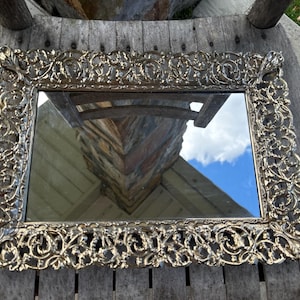 Op de afbeelding: Een sierlijke spiegel met een decoratief, bloemmotief frame, afgewerkt in zilver. De spiegel reflecteert een houten oppervlak en een blauwe lucht.