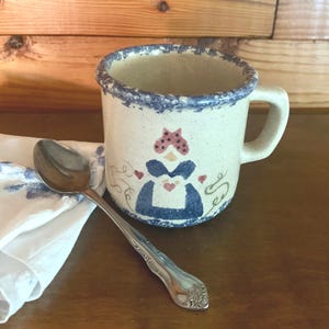 Może przedstawiać: Biały kubek ceramiczny z malowanym wzorem w kolorze niebieskim i różowym przedstawiającym kobietę w niebieskiej sukience i czerwono-białym nakryciu głowy w kropki. Kubek ma niebieski rant i uchwyt.