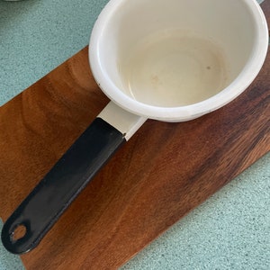 May include: A small, white enamel saucepan with a black handle rests on a wooden cutting board. The saucepan has a rounded shape and a single handle. The handle is black with a white stripe near the pan. The cutting board is a dark brown color.