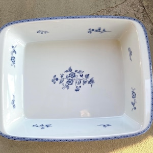 Puede incluir: Un plato para hornear de cerámica blanca con un patrón floral azul alrededor del borde. El plato es rectangular y tiene un borde ligeramente elevado.