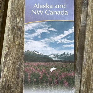 May include: A folded travel map for Alaska and NW Canada, featuring a scenic landscape of mountains, a field of pink flowers, and a blue sky with clouds. The map is from AAA/CAA.