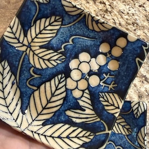May include: A square ceramic dish featuring a deep blue glazed background with a cream-colored botanical pattern of leaves and clusters of small round berries. The design includes dark outlines defining the foliage and fruit across the surface.