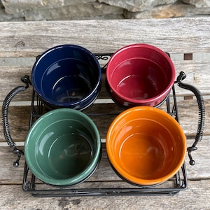 May include: A set of four round ceramic bowls in navy blue, deep red, sage green, and bright orange, each featuring a speckled finish. The bowls are arranged in a rectangular black wire metal caddy that includes two side handles with coiled wire.
