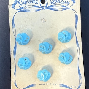 May include: A vintage card of six light blue plastic buttons. The buttons have a floral design and are 24-5/8 inches in size. The card reads "Supreme Quality" and "Made in U.S.A."