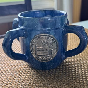 May include: A blue ceramic mug with two handles and a silver emblem. The mug has a glossy finish and a decorative emblem with an image of a building. The mug is sitting on a woven surface.
