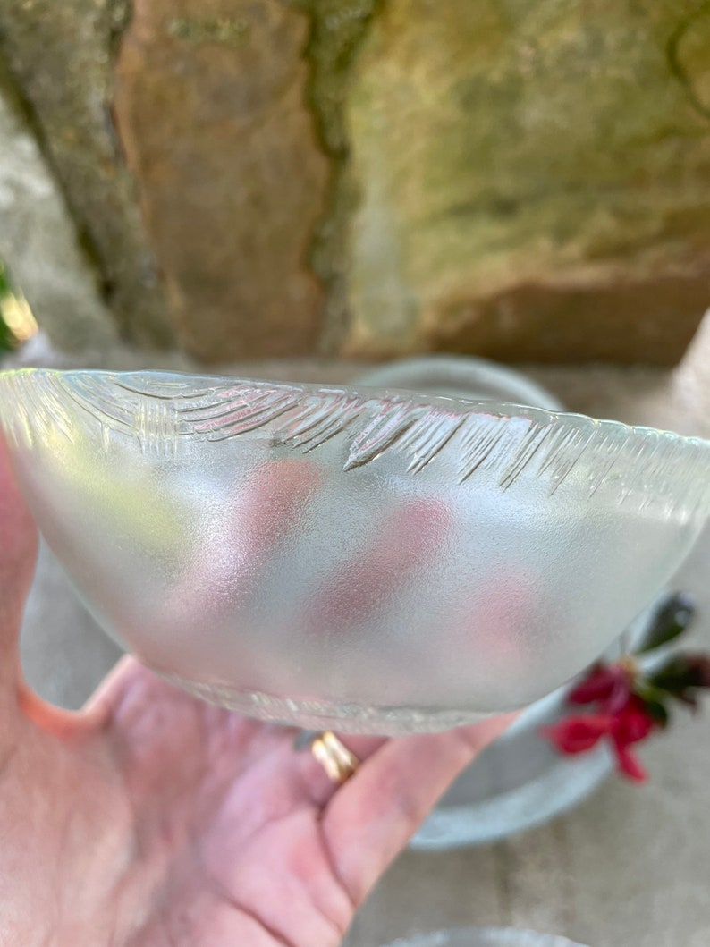 May include: A frosted glass bowl with a textured rim. The bowl is held in a hand, with a blurred background of stone and foliage. The bowl's interior reflects soft pink and yellow hues.