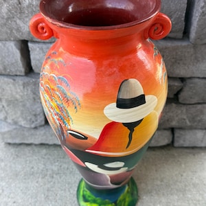 Puede incluir: Jarrón de cerámica pintado a mano con una vibrante escena de atardecer. El jarrón tiene una parte superior ancha y abierta con dos asas pequeñas y una base de pedestal. La obra de arte representa a una persona con sombrero, palmeras y un paisaje colorido. El jarrón mide unos 25 cm de alto.