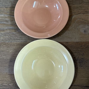 May include: Two ceramic bowls, one peach and one pale yellow, with a slightly raised rim. The bowls are empty and sitting on a wooden surface.