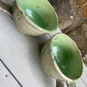 May include: Two green ceramic mugs with white and brown speckled glaze. The mugs have a rounded shape and a handle.