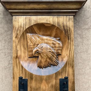 May include: A wall-mounted wooden shelf with a carved eagle head and an American flag in a circular recessed center. The piece features a dark brown wood grain finish and two black metal double hooks attached to the lower front section.